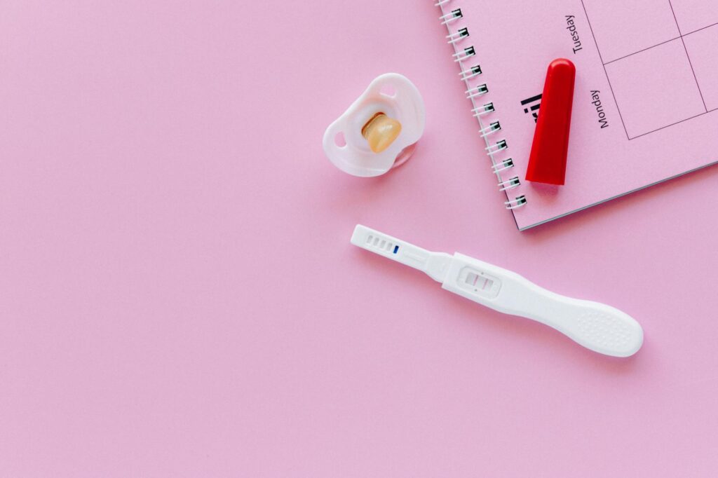 A positive pregnancy test on a pink surface with a pacifier and planner.